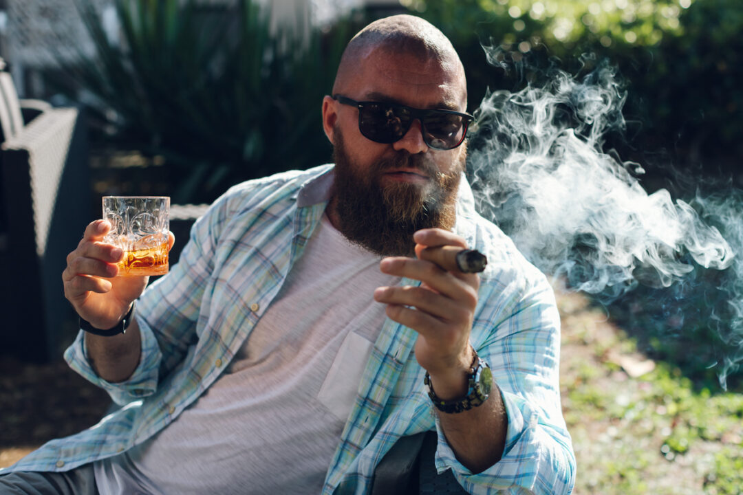 Portrait of an attractive bearded business man smoking a cigar with glass of whiskey in a summer cafe.